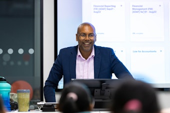 An instructor smiles engagingly while presenting information, standing behind a desk in a modern classroom environment with a screen display.