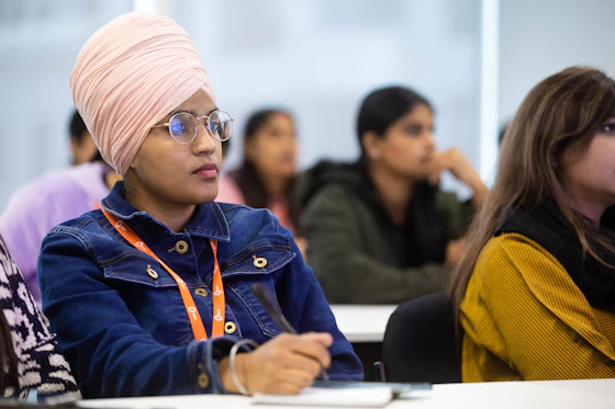 A student in a lecture hall is actively taking notes with a focused gaze towards the front of the class.