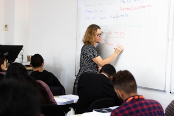 A female instructor writing "Answer Structure" on a whiteboard. The focus is on the teaching action, with the backs of students visible in the foreground.