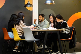 A diverse group of four students sitting around a high table in a social study area, actively discussing materials with open books and animated gestures.