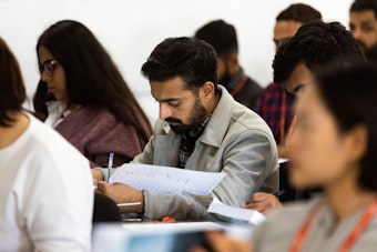 A male student seated at a desk in a lecture hall, concentrating intensely on writing notes or an assignment on a sheet of paper.