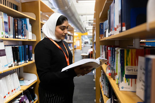 A student browsing and reading a book in the library stacks. A student standing between library shelves, holding an open book and reading intently while looking at the text.