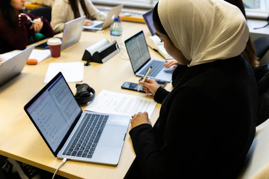 Students working together on laptops and papers at a shared table in a modern study space. Students gathered around a table with multiple laptops and study materials, actively working and taking notes in a group setting.