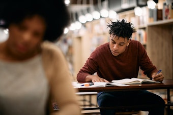 A male student seated at a library desk writing in a notebook, with bookshelves and blurred study lights in the background.