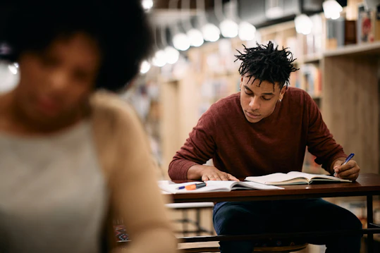 A male student seated at a library desk writing in a notebook, with bookshelves and blurred study lights in the background.