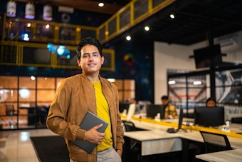 A man holding a tablet and smiling, standing in a large, modern office space with multiple workstations visible behind him.