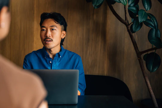 A man with a blue shirt sitting opposite another person (out of focus), speaking with an earnest expression during a meeting. A laptop is visible on the dark table.