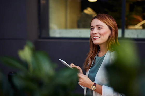 A woman with red hair, smiling widely, stands outside a building holding a tablet in her hands.
