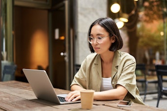A woman with short hair and glasses working on a silver laptop at an outdoor table with a paper cup and phone nearby.