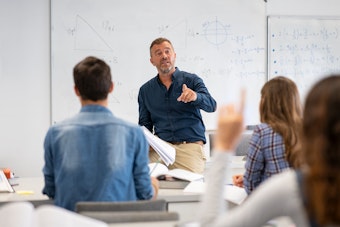 A male instructor pointing and speaking to students in a classroom. A student in the foreground has her hand raised, ready to answer.