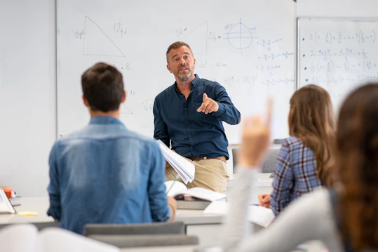 An instructor actively directs a question to the class, encouraging student participation. A male instructor pointing and speaking to students in a classroom. A student in the foreground has her hand raised, ready to answer.