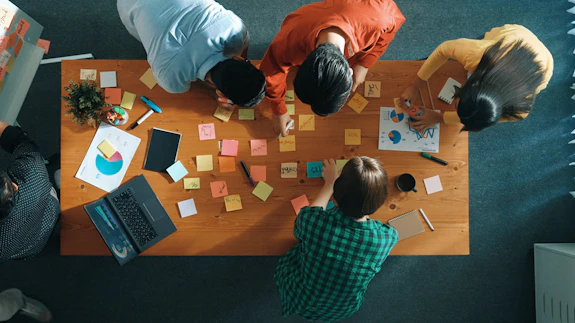 An overhead shot of a diverse team of five people leaning over a wooden table covered in colorful sticky notes, a laptop, charts, and notepads, actively collaborating during a brainstorming session.