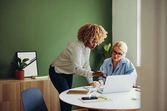 Two professional women are seated and standing at a small round table with a laptop. One woman with blonde hair and glasses is seated looking at the screen, while another woman with curly blonde/brown hair is standing, pointing at the laptop screen and discussing the content.
