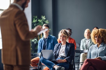 A woman in a dark blazer and jeans is seated in an audience during a presentation, smiling and looking toward the out-of-focus male speaker in a brown suit. She has a laptop resting on her lap, and other diverse attendees are visible in the background.