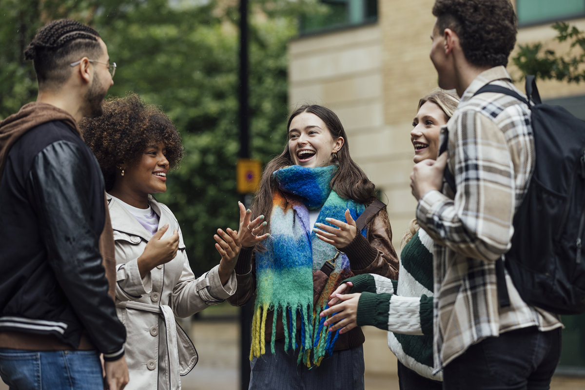 A diverse group of five students is standing outside in an urban setting, smiling and laughing while having an engaging conversation.