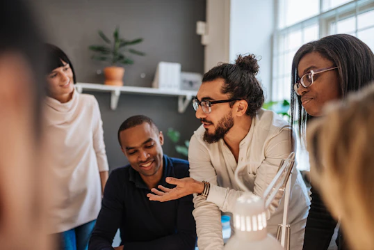 A close-up, indoor shot of a diverse group of business professionals gathered around a table. A man with a beard and glasses is actively speaking and gesturing with his hand to his colleagues.