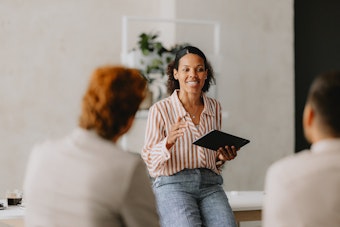 A smiling woman in a striped shirt and patterned pants is seated on a desk, holding a black tablet and gesturing while speaking to colleagues who are out of focus in the foreground, leading a discussion or presentation in a bright office.