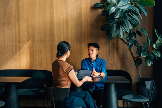 Two people are seated across from each other at a dark table in a modern space with wood-paneled walls and a large plant. A woman is showing content on a tablet to a man who is attentively listening.