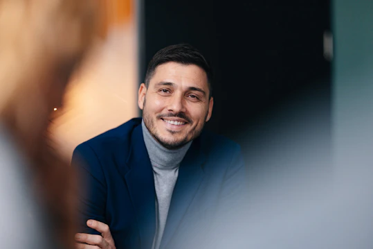 A close-up portrait of a man in a blue blazer and grey turtleneck sweater, smiling genuinely at the camera during a business meeting. The foreground and background are blurred.