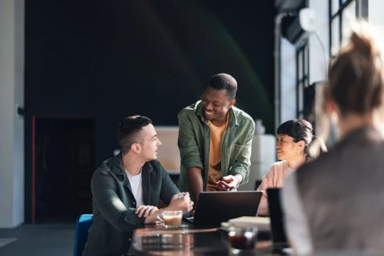A diverse group of three colleagues is gathered around a laptop on a table in an office. A smiling Black man is standing and talking to a seated man and a seated Asian woman, actively engaged in a discussion.