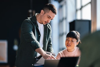 A man is standing and pointing at a notebook held by a seated woman, who is looking intently at him, while a laptop is visible in the foreground, suggesting close collaboration on a task.