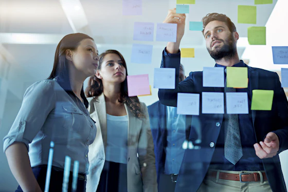 A low-angle shot of a business team of three people (two women and one man) standing in an office and looking through a clear glass board covered with small, colorful sticky notes during a strategy session. The man is placing a note higher up on the board.