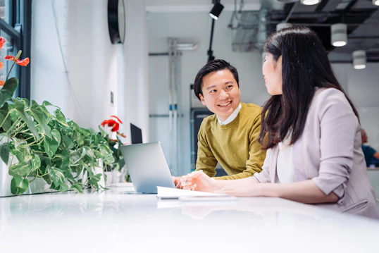 A man and a woman are seated across from each other at a white desk in a modern office, engaged in a conversation. A laptop is open on the table between them, and indoor plants are visible in the foreground.
