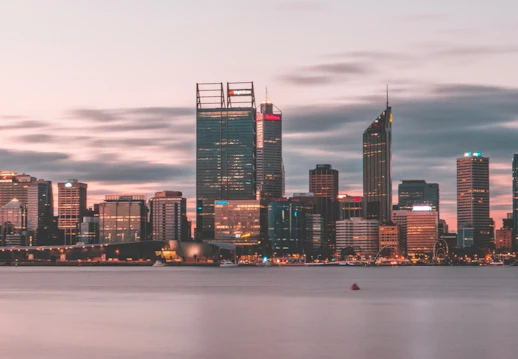 A city skyline of Perth in the early evening, with a calm river and tall skyscrapers lit up