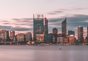 A city skyline of Perth in the early evening, with a calm river and tall skyscrapers lit up