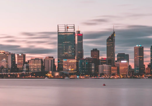 A city skyline of Perth in the early evening, with a calm river and tall skyscrapers lit up